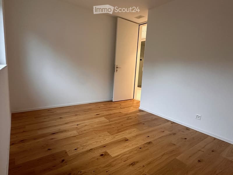 Empty room with light wooden flooring and white walls, featuring a partially open door leading to another space.
