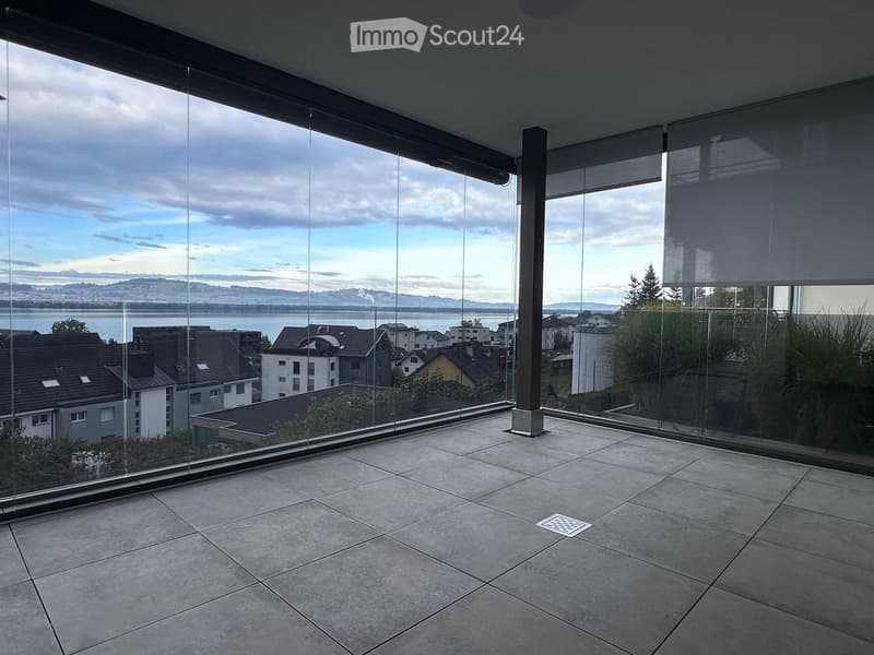 Modern balcony with large glass walls, tiled floor, and a view of a lake and surrounding buildings.