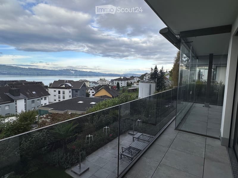 Modern balcony with glass railing, tiled floor, small garden area with plants, and outdoor furniture including lounge chairs and a small table.