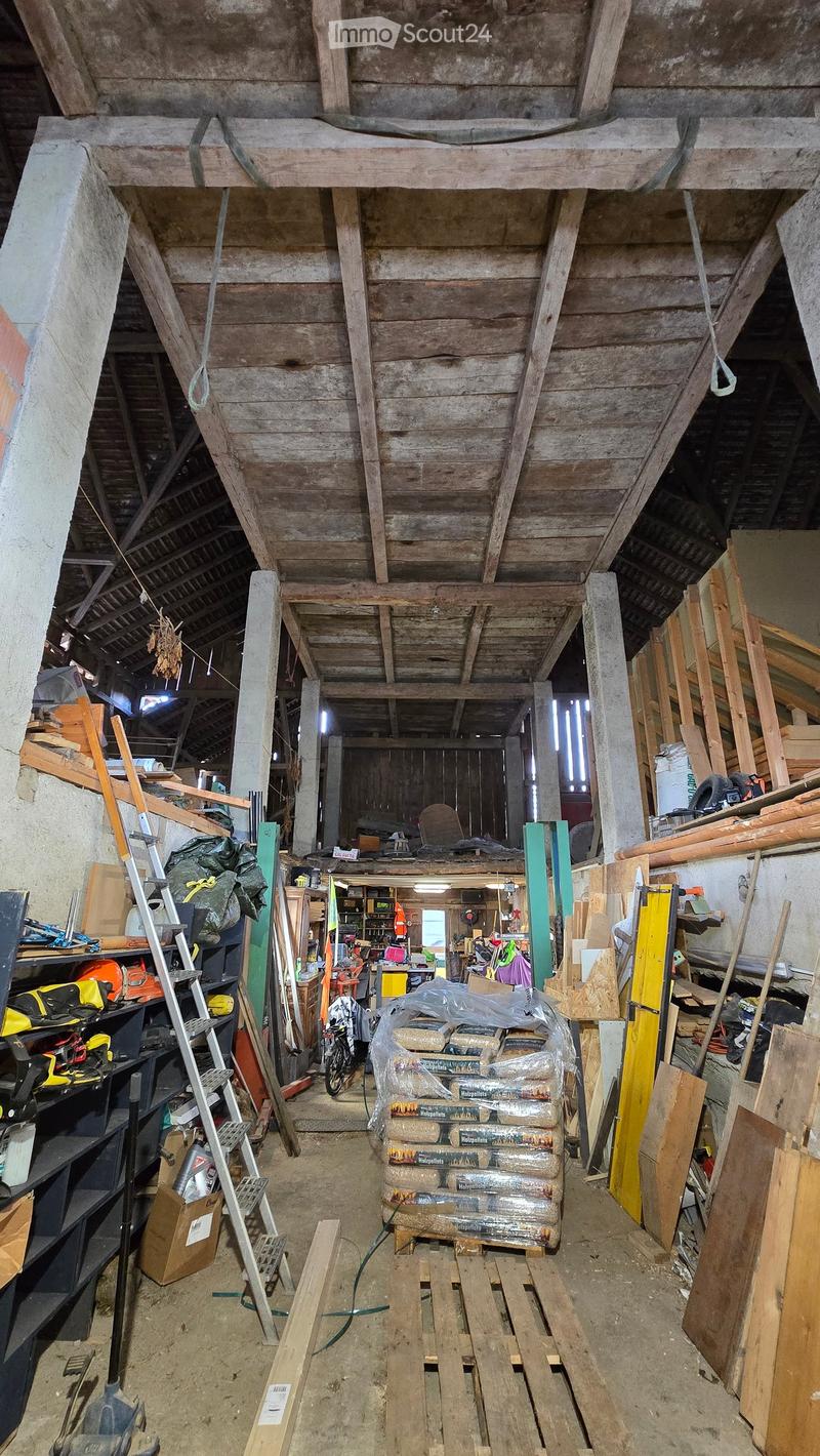 The image shows an unfinished attic or storage space with exposed wooden beams and a high, slanted wooden ceiling. The floor is concrete, and the area is filled with various construction materials, tools, and storage items, including wooden planks, plasti