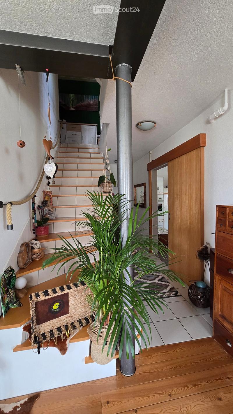 Staircase leading to an upper level, with wooden railings, a large potted plant in the foreground, and decorative items on shelves along the wall.