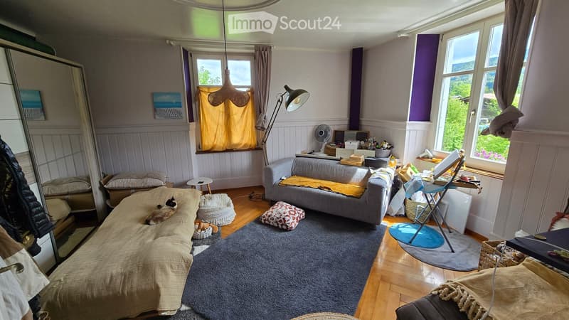 Cozy living room with a grey sofa, a cat bed, a small table, a fan, a mirror, a lamp, and a stool. The room features wooden flooring, large windows with curtains, and a view of greenery outside.