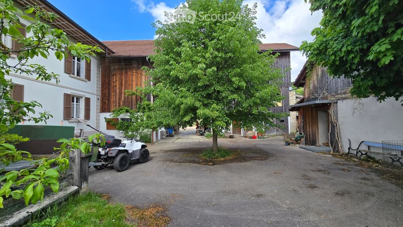 A rural property with a two-story house, wooden barn, and surrounding greenery. A white ATV is parked near the house, and there are outdoor seating areas and trees.