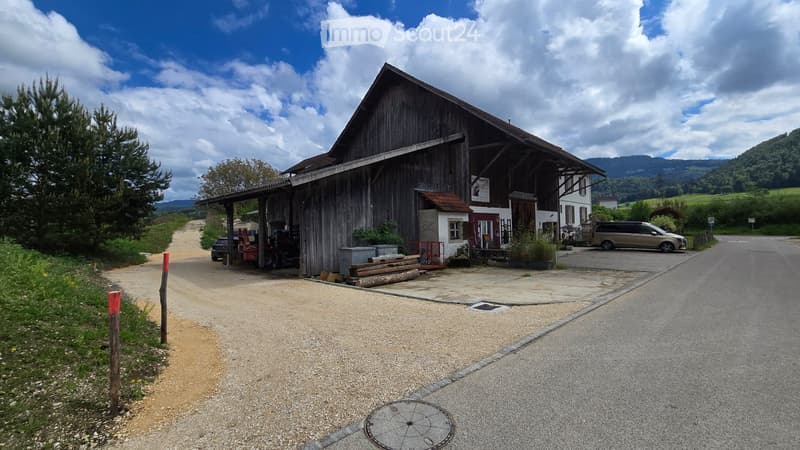 A large wooden barn with a covered entrance, surrounded by a gravel driveway and parked vehicles, with a scenic rural backdrop.