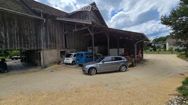 Large wooden barn with an open garage area, several parked cars including a grey sedan, a blue van, and a white car, surrounded by a gravel driveway and rural landscape.