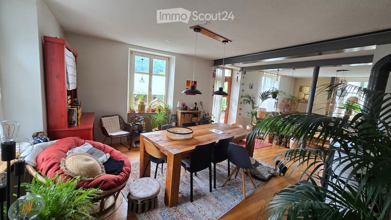 Bright, open living and dining area with wooden floors, large windows, and modern decor. Features a red sofa, wooden dining table with chairs, hanging pendant lights, and various plants.