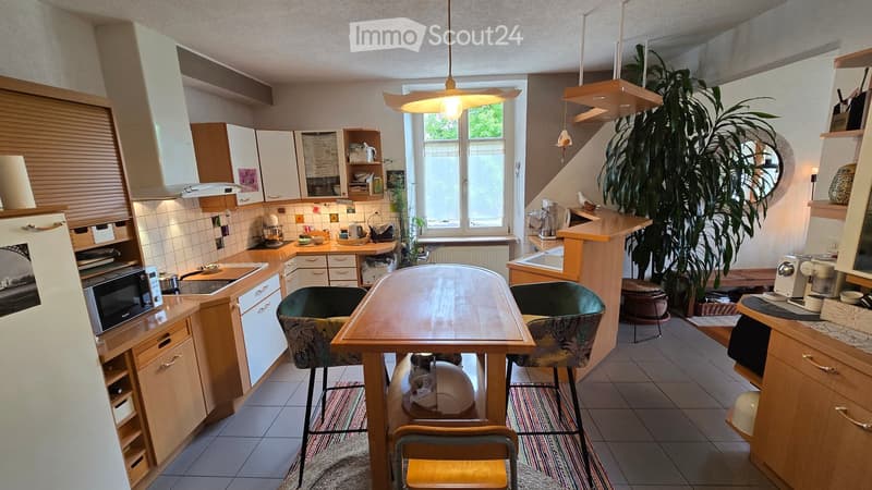 Bright kitchen with wooden cabinets, a central dining table with chairs, and various appliances including a refrigerator, microwave, and oven. A large window provides natural light, and a potted plant adds a touch of greenery.