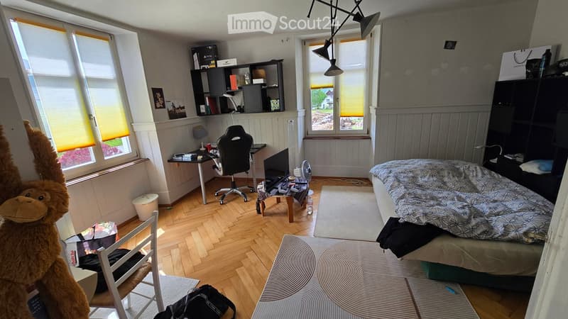 A well-lit bedroom with wooden flooring, a bed with a patterned duvet, a desk with a chair, shelves, and a fan. The room features large windows with blinds and a cozy atmosphere.