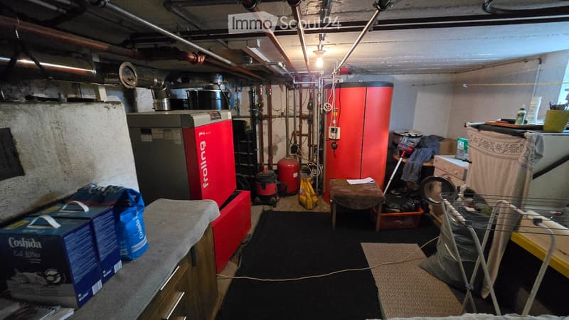 The image shows a basement utility room with a washing machine, dryer, furnace, and various storage items. There are also a few stools, a small table, and a basket visible.