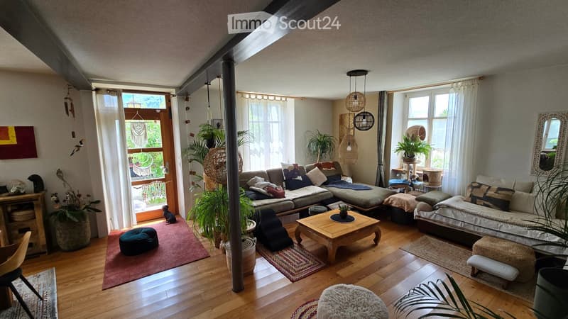 A cozy living room with wooden flooring, large windows, and a mix of modern and bohemian decor. Features include a sectional sofa, coffee table, ottoman, hanging pendant lights, and several potted plants.