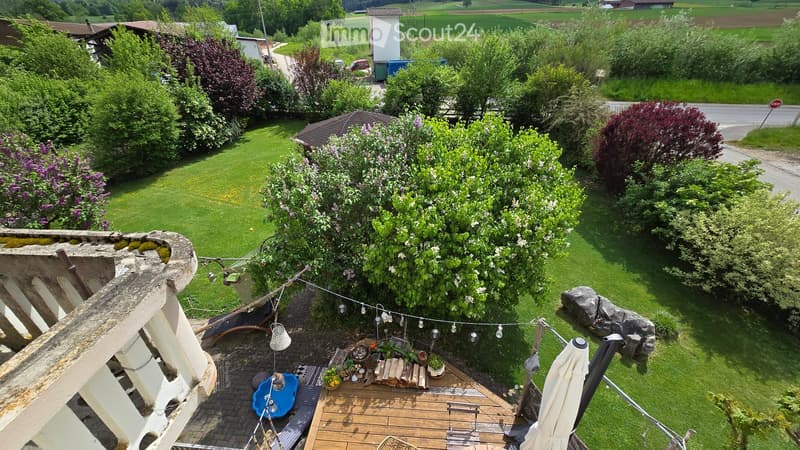 A well-maintained garden with a wooden deck, outdoor furniture, potted plants, and a small table with chairs. The area is surrounded by lush greenery and features a string of lights for ambiance.