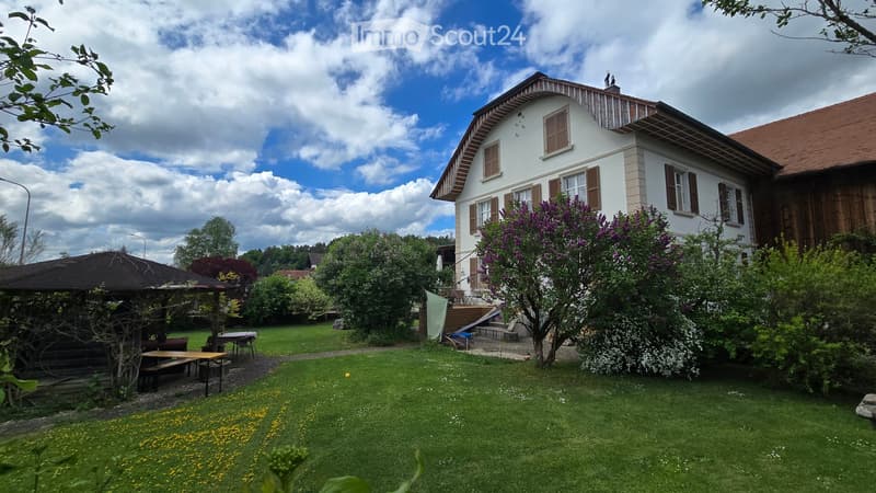 Lush garden with a two-story house in the background, featuring a well-maintained lawn, colorful flowers, and a wooden pergola with outdoor seating.
