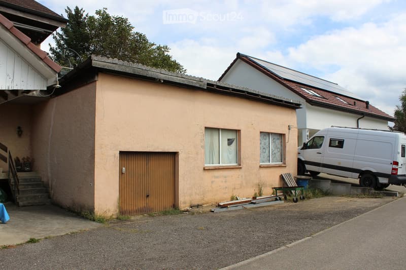 Small house with a garage, two windows, and a tree nearby