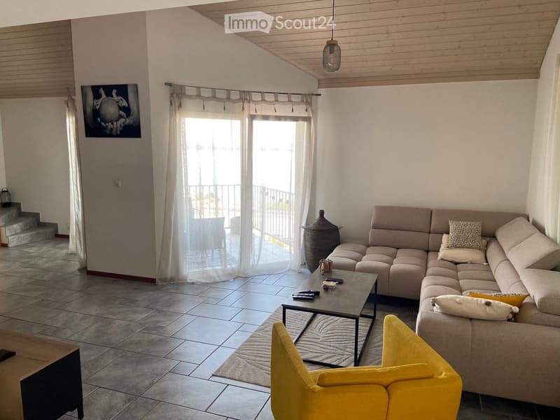 Modern living room with a large sectional sofa, coffee table, yellow armchair, and a view to a balcony. The room features tiled flooring, white walls, and a hanging pendant light.