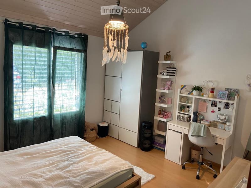 Cozy bedroom with a bed, wardrobe, desk, chair, shelves, and decorative items. Features a hanging feather lamp and a large window with dark curtains.