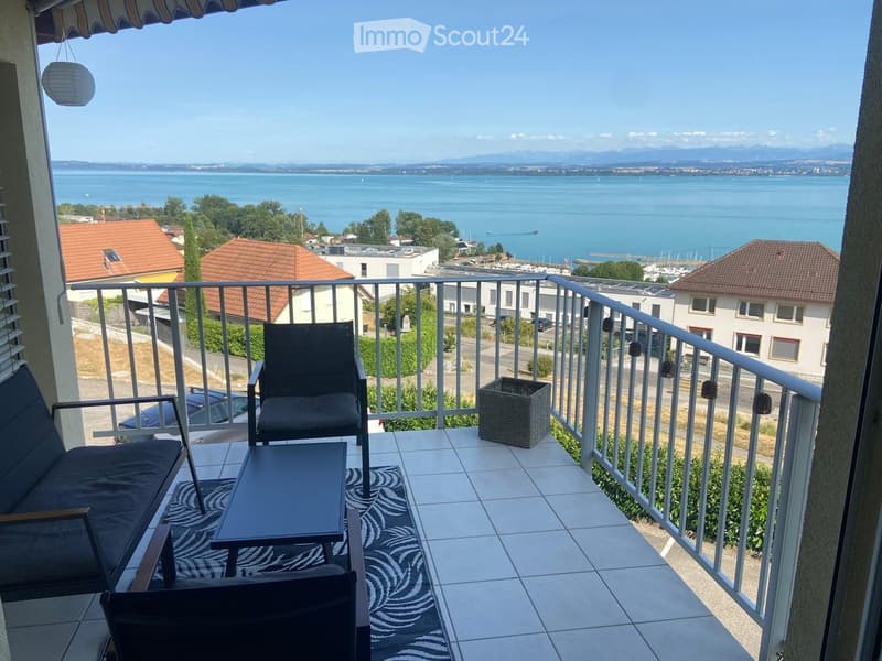 Balcony with a scenic view of a lake and distant mountains, furnished with a sofa, chairs, a table, and a wicker basket.