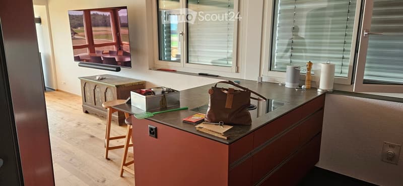 Modern kitchen with wooden flooring, a countertop, and various items including a handbag, paper towel, pepper grinder, and a screen. There is a small table with a stool and a trash bin nearby.