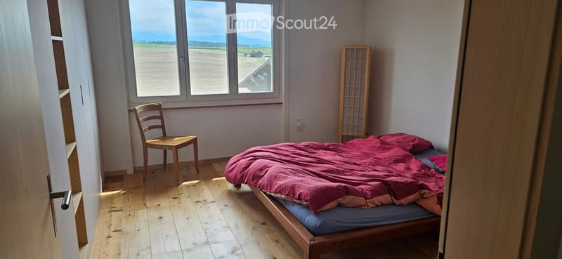 Cozy bedroom with wooden floor, large windows overlooking a field, a bed with red bedding, a wooden chair, and a screen.