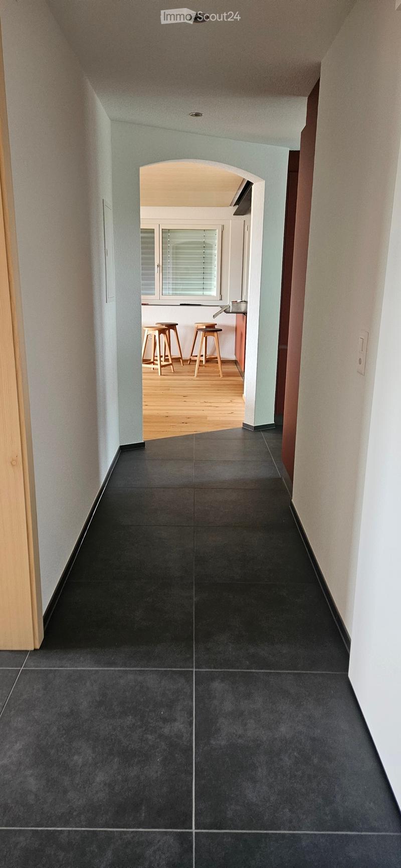 A modern hallway with dark grey tiled flooring, light grey walls, and an arched opening leading to a room with wooden stools and a window.