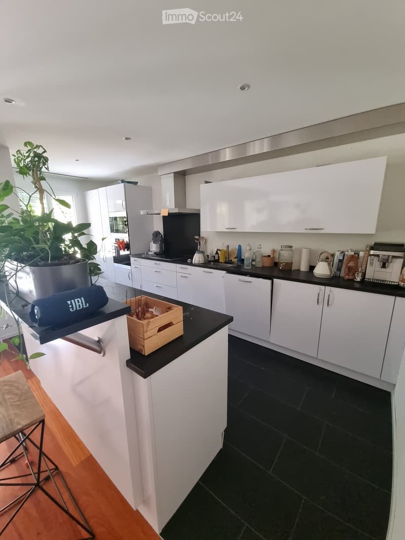 Modern kitchen with white cabinets, black countertops, and various appliances including a coffee maker and teapot. A large plant and wooden crate are on the counter.
