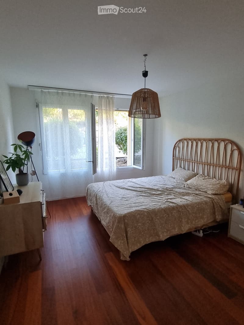Bedroom with wooden flooring, a bed with a decorative headboard, a nightstand with a lamp, a plant, and a window with sheer curtains.