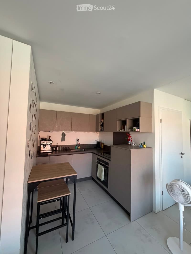 Modern kitchen with light grey cabinets, a wooden dining table with two stools, a built-in oven, a sink with a faucet, and a fan on the floor.