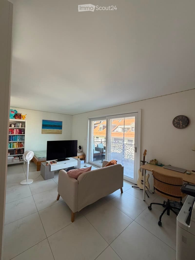 A modern living room with a sofa, coffee table, TV, bookshelf, and a desk area with a laptop and guitar. The room features large windows and a sliding glass door leading to a balcony.