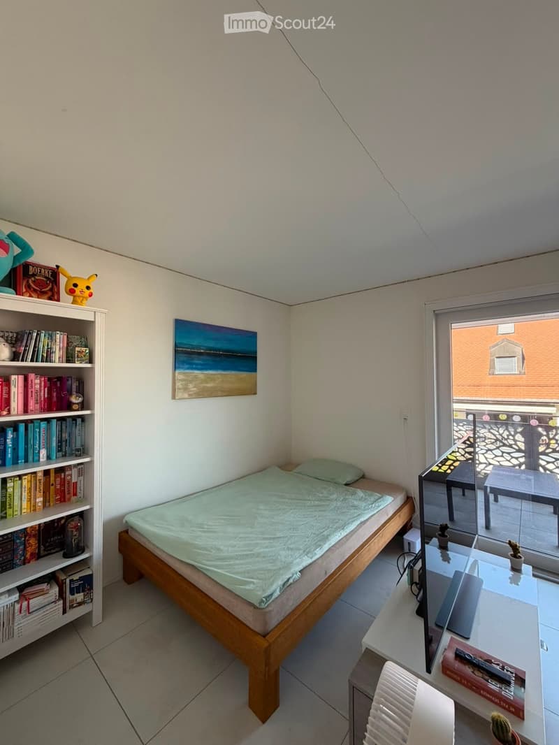 A small, well-lit bedroom with a wooden bed, a bookshelf filled with books and decorative items, a desk with a screen and chair, and a painting on the wall. The room has a view of a balcony area through a large window.