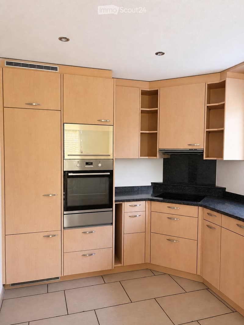 Modern kitchen with light wood cabinets, stainless steel oven, black countertop, and tiled floor.