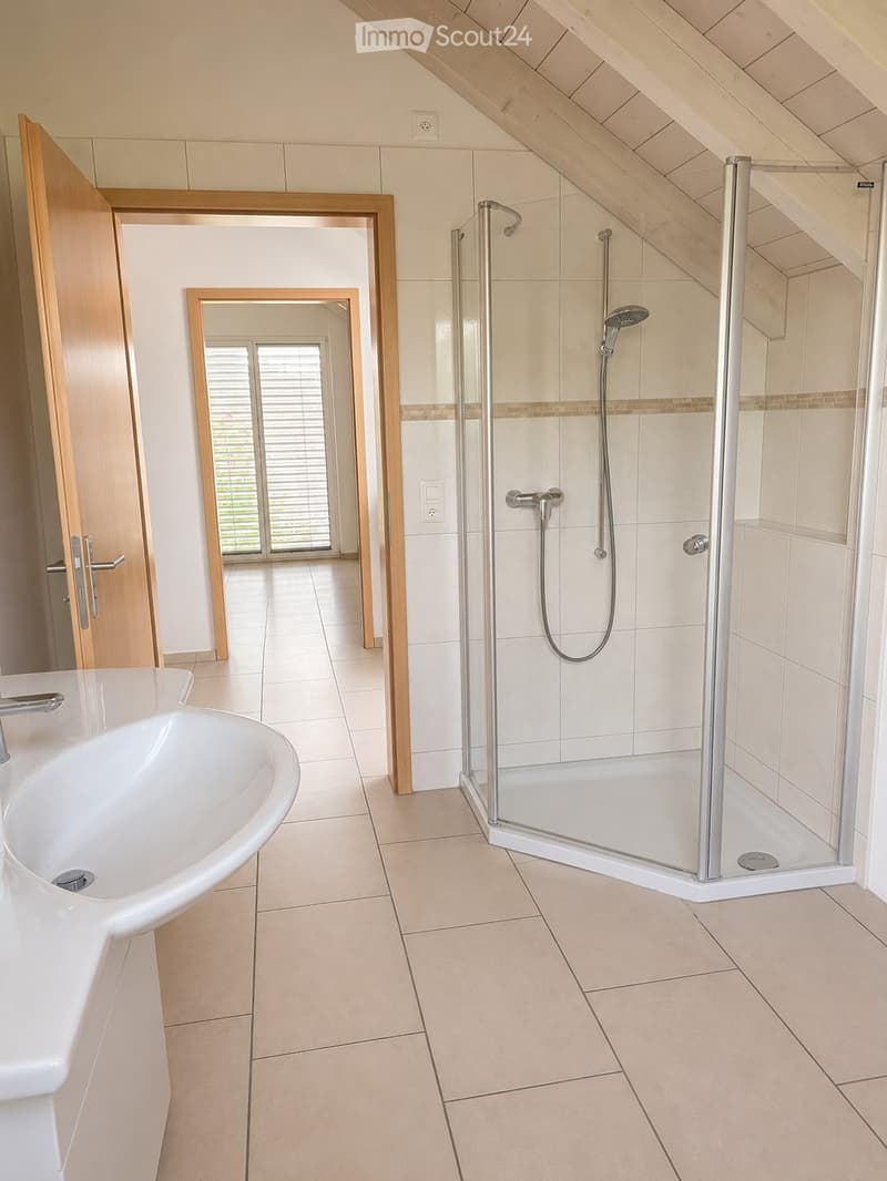 Modern bathroom with a white sink, glass-enclosed shower, and a large mirror. The room features beige tiles and wooden trim.