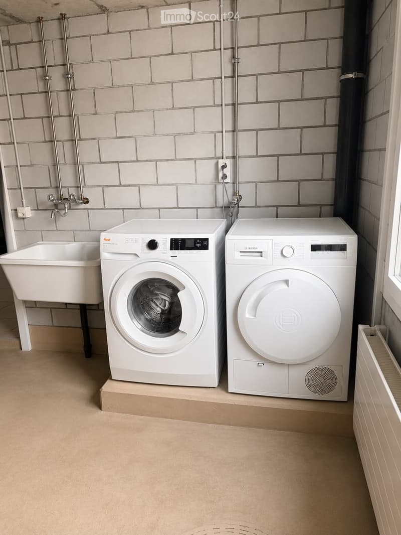 A small bathroom with a washing machine, dishwasher, and a sink, featuring white tiled walls and a beige floor.
