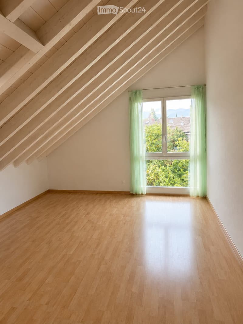 Empty attic room with wooden flooring, slanted wooden ceiling beams, and large window with green curtains, offering a view of greenery and rooftops.