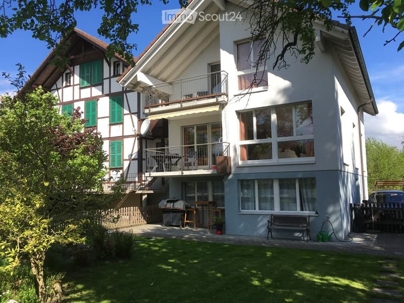 Two-story house with white walls, green shutters, and a balcony. The house features a well-maintained garden with a lawn, a bench, and outdoor furniture.