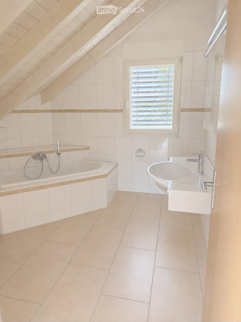 Modern bathroom with a bathtub, sink, and toilet paper holder. The room features white tiles, a window with blinds, and a sloped wooden ceiling.