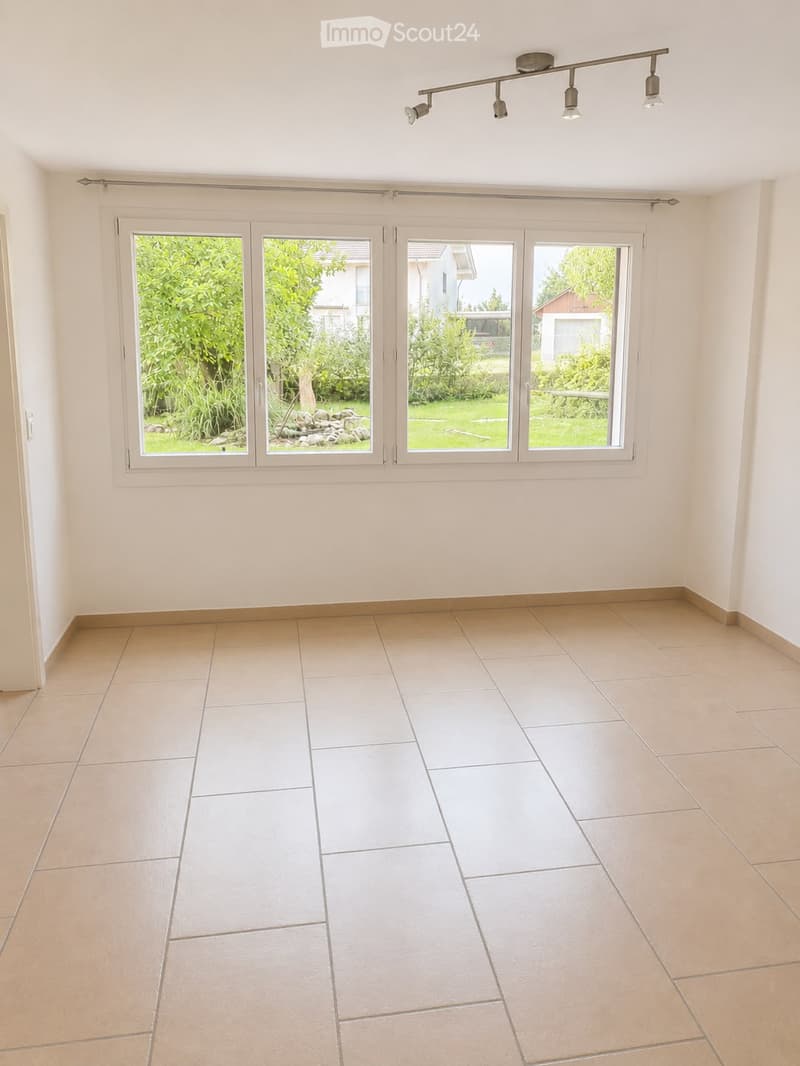 Empty room with beige tiled flooring, large sliding glass doors leading to a garden, and recessed lighting fixtures on the ceiling.