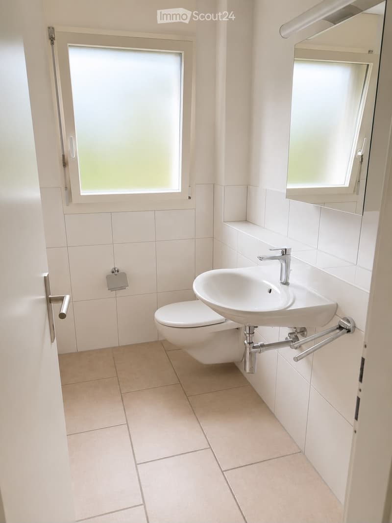 Modern bathroom with white tiles, a toilet, a sink with a modern faucet, and a mirror above the sink. The room has frosted glass windows and a beige tiled floor.