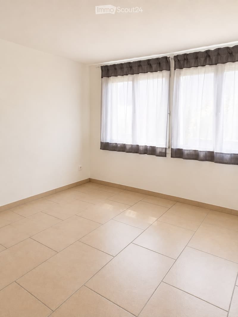 Empty room with beige tiled flooring and white walls, featuring a large window with white and dark grey curtains.