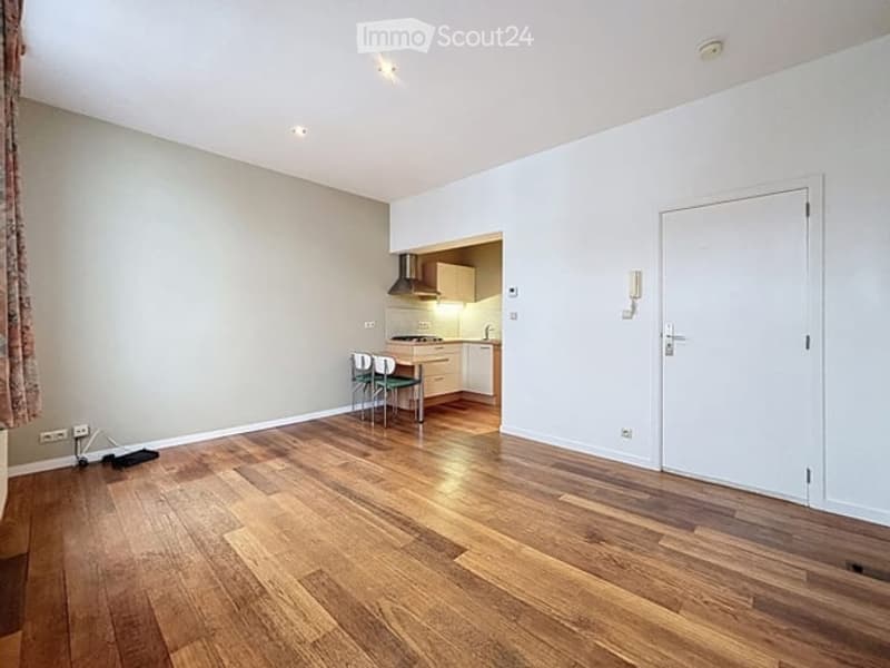 Bright, open-plan living and dining area with wooden flooring, white walls, and a small kitchen area. Includes a dining table with chairs, kitchen cabinets, and a stove with a range hood.