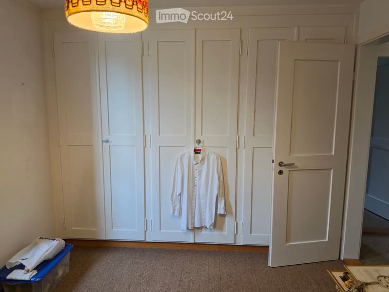 A small, unfurnished room with a white wardrobe, a shirt hanging on the door, and a storage box on the floor.