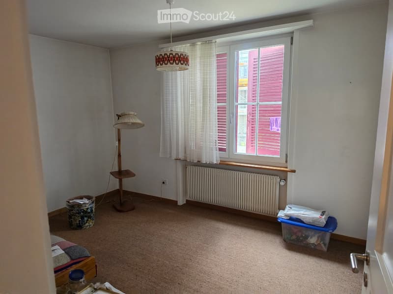 Empty room with beige carpet, white walls, large window with white blinds, radiator, floor lamp, and a few storage items.