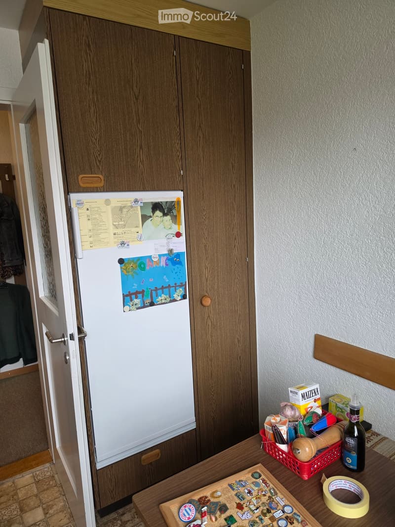 A small hallway with a refrigerator covered in magnets and drawings, a wooden cabinet, and a table with various items including a bottle, tape, and a basket.