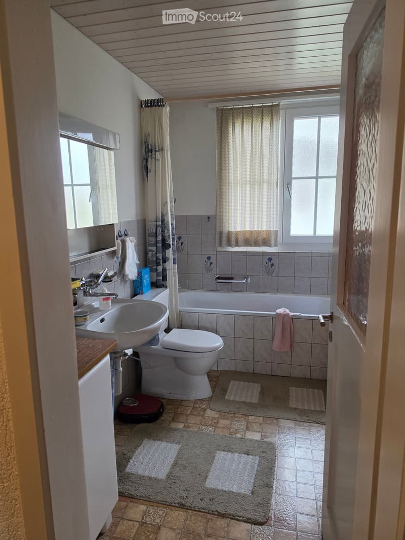 Bathroom with a sink, toilet, bathtub, and mirror. The room features tiled walls and floors, with a window and curtains providing natural light.