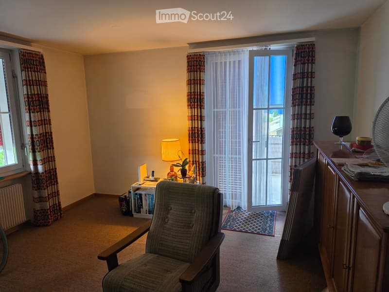 A cozy living room with a patterned armchair, a small table with a lamp, a cabinet with a fan and cup, and a window with curtains leading to a balcony.