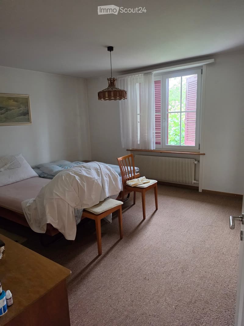 Bedroom with a bed, two chairs, a small table, a radiator, and a picture frame on the wall. The room has carpeted flooring and large windows with white curtains.