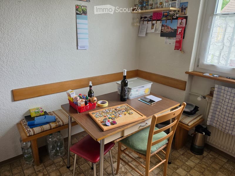 A small, cozy room with a wooden table, two chairs, a shelf with various items, and a calendar on the wall. The table is cluttered with a bottle, a plastic container, and a puzzle board.