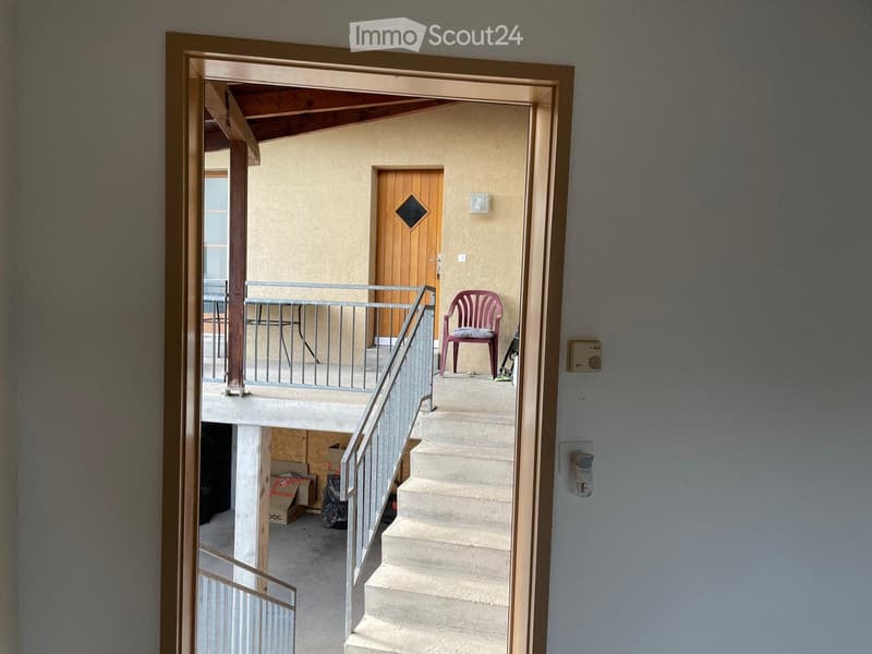 A narrow hallway with a staircase leading upwards, featuring a metal railing, a wooden door at the end, and a small red chair on the landing.