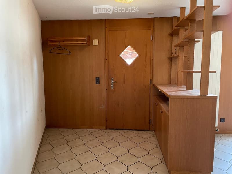 A narrow hallway with wooden doors, built-in wooden shelving, and a coat rack. The floor is tiled with hexagonal tiles.