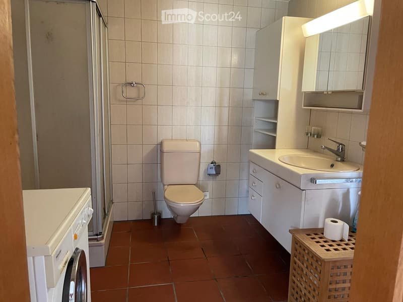 A modern bathroom with beige tiled walls, a toilet, a sink with a mirror cabinet, a washing machine, and a wooden basket holding toilet paper. The floor is tiled in a warm brown color.