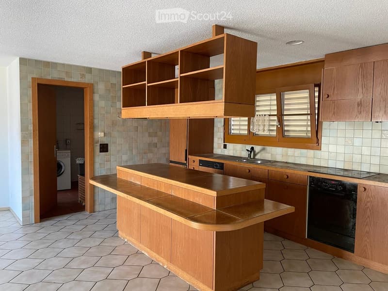 Wooden kitchen with built-in cabinets, a central island counter, sink, and oven. Open shelving above the counter, tiled backsplash, and a washing machine visible in the adjacent room.