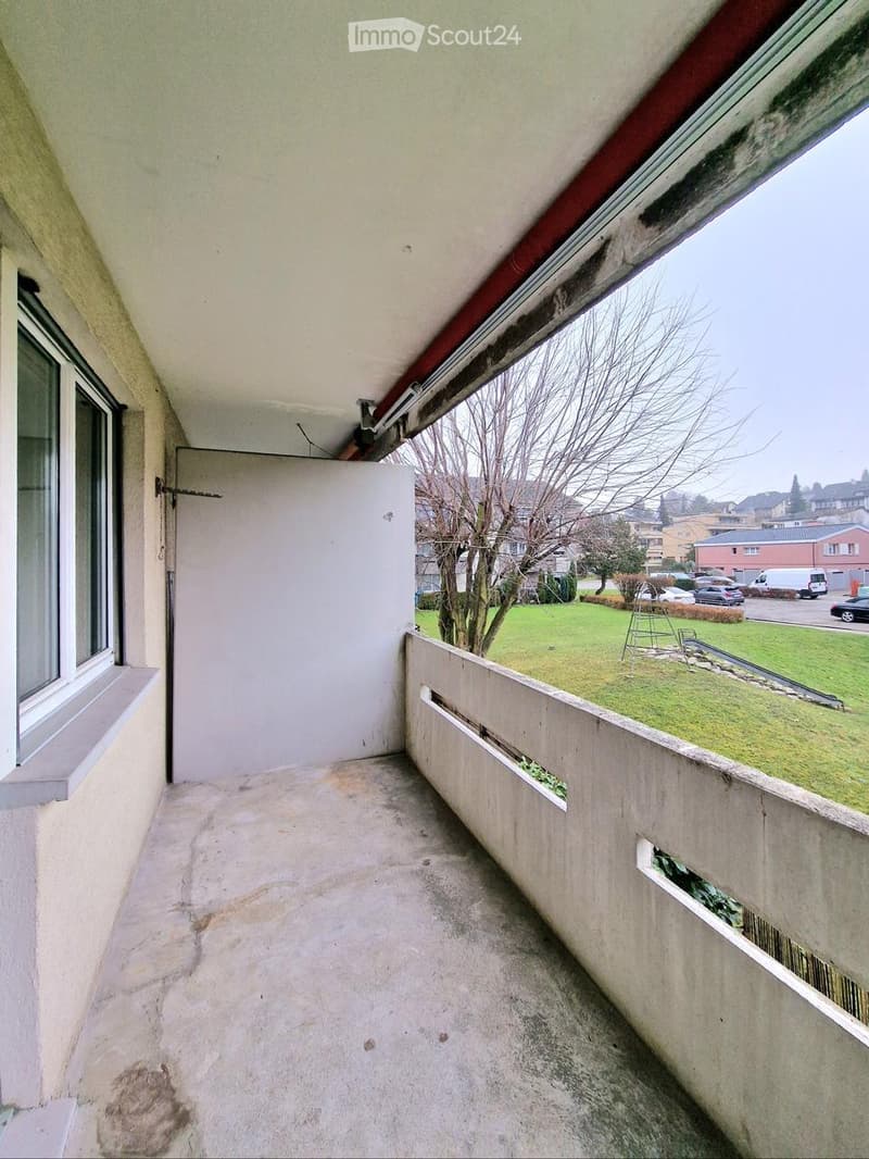 Balcony with concrete floor, white walls, and a door leading to the interior. The view shows a green lawn, trees, and parked cars in the background.